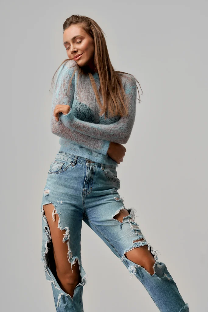 Frau in hellblauem, transparentem Strickpullover trägt hellblaue ripped jeans mit großen seitlichen Rissen und posiert vor grauem Studiohintergrund.