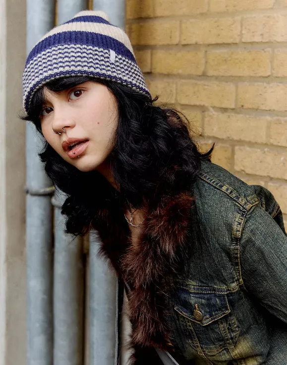 Person mit dunklem, welligem Haar trägt eine blau gestreifte Strickmütze und eine Jeansjacke mit Fellkragen vor einer gelben Backsteinwand.