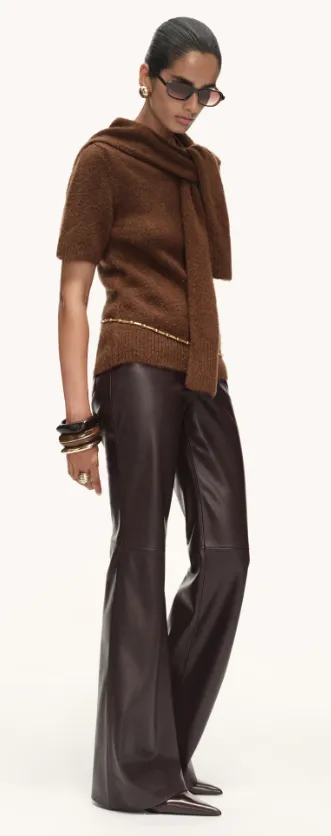 Moderne dunkelbraune Lederhose in Kombination mit einem braunen Strickoberteil und spitzen Schuhen, getragen von einer stehenden Person im Studio.