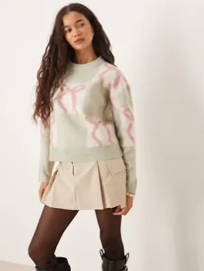 Damen-Weihnachtspullover in Salbeigrün mit rosa Geschenkband-Muster, kombiniert mit kurzem Faltenrock und Strumpfhose.