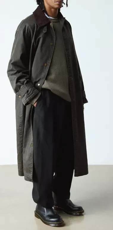 Herrenoutfit mit schwarzen Chelsea Boots, weiter Stoffhose und langem Mantel in dunklen Tönen.