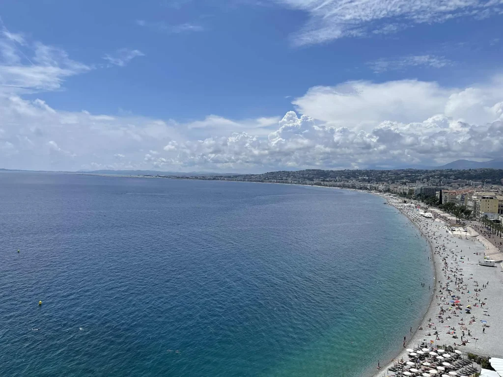 Weite Strandbucht an der Côte d’Azur mit klarem blauem Wasser, heller Promenade und belebtem Kiesstrand in Nizza – ideales Ziel für spontane Last-Minute-Reisen Richtung Süden.