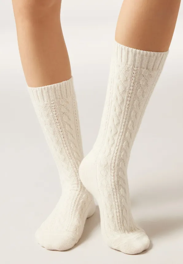 Cremefarbene, kniehohe Stricksocken mit feinem Zopfmuster an den Beinen einer stehenden Person.