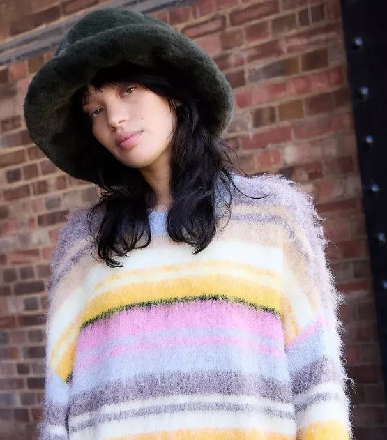 Person mit dunklem Haar trägt einen flauschigen, olivgrünen Bucket-Hut aus Kunstfell und einen weichen, gestreiften Pullover vor einer Backsteinwand.