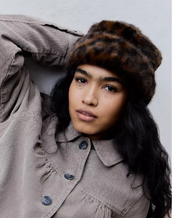 Frau mit dunklem Haar trägt einen flauschigen Pillbox-Hut mit Leopardenmuster und eine hellbraune Cordjacke.