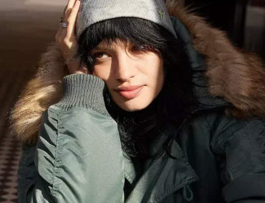 Person mit dunklem Haar trägt eine graue, leicht gemusterte Mütze und eine grüne Winterjacke mit Fellkapuze, während sie in der Sonne sitzt.