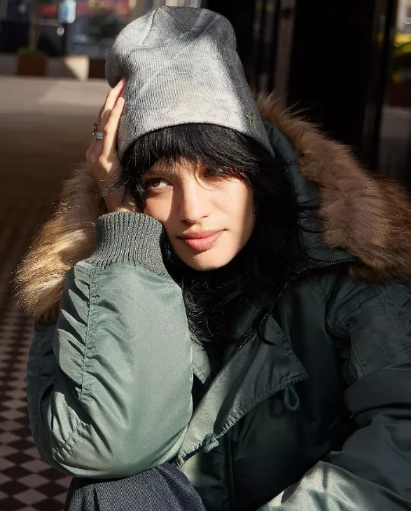 Person mit dunklem Haar trägt eine graue, leicht gemusterte Mütze und eine grüne Winterjacke mit Fellkapuze, während sie in der Sonne sitzt.