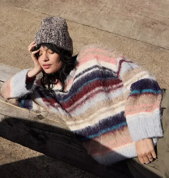 Person mit dunklem Haar trägt einen grob gestrickten Beanie und einen gestreiften, flauschigen Pullover, während sie entspannt auf einer Holzbank in der Sonne liegt.