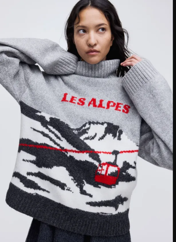 Damen-Weihnachtspullover in Grau mit alpinem Motiv, roter Seilbahn und „Les Alpes“-Schriftzug, getragen in einem modernen Winterlook.