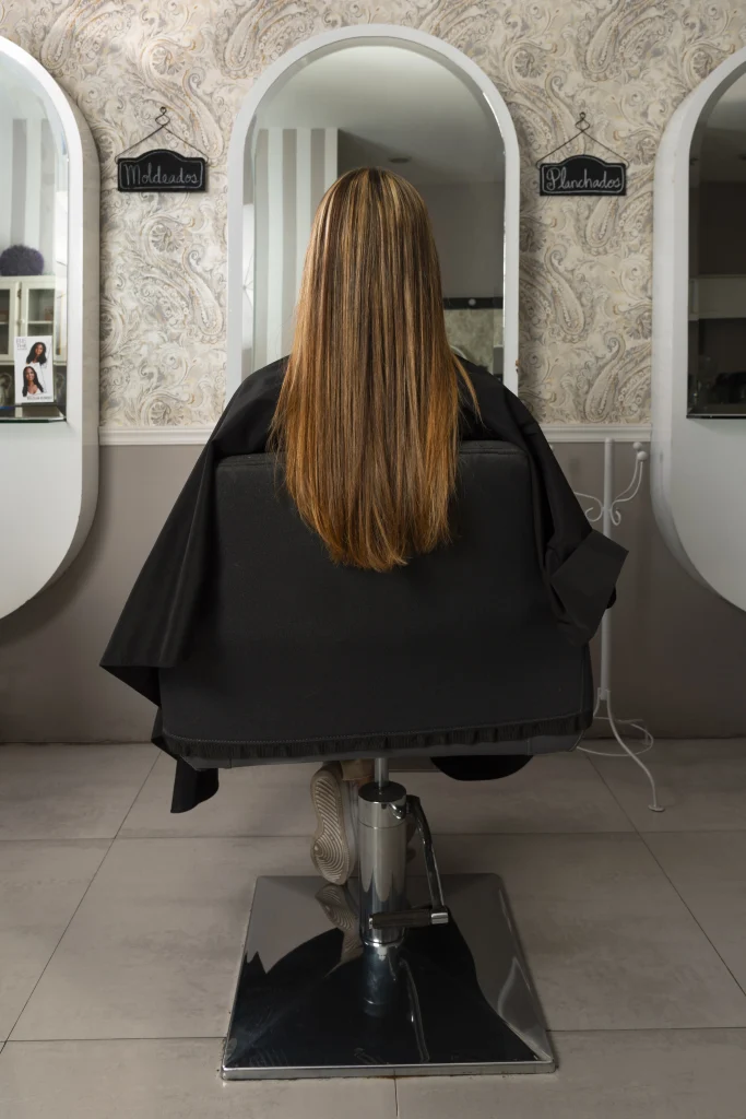 Frau mit langen, glatten Haaren sitzt im Friseursalon auf einem Stuhl, von hinten fotografiert, während sie auf ihre Behandlung wartet.