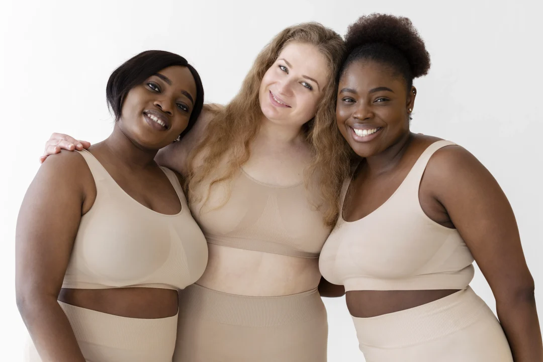 Drei Frauen verschiedener Körperformen tragen hautfarbene Shapewear von Creamy Fabrics und stehen lächelnd Arm in Arm vor hellem Hintergrund.