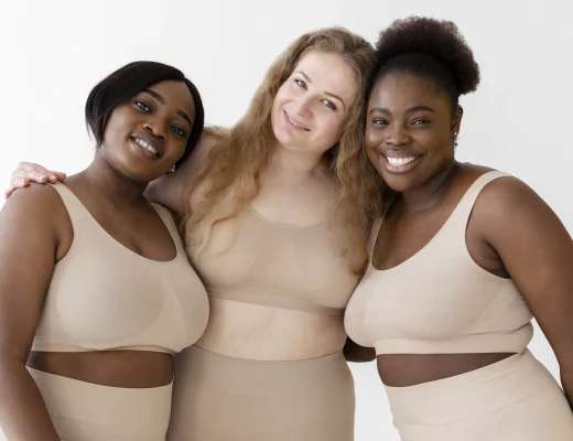 Drei Frauen verschiedener Körperformen tragen hautfarbene Shapewear von Creamy Fabrics und stehen lächelnd Arm in Arm vor hellem Hintergrund.