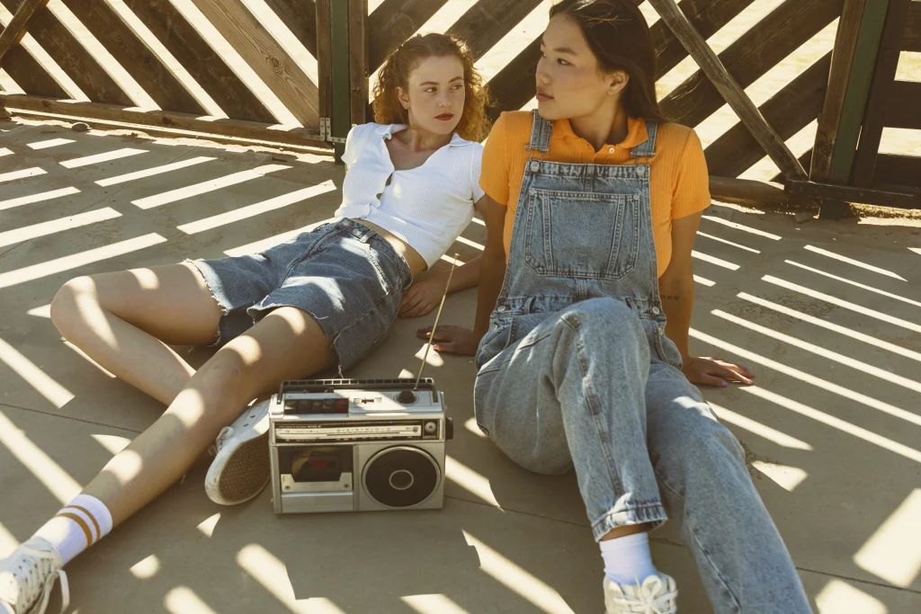 Zwei junge Frauen im Y2K Look mit Jeans-Shorts und Latzhose sitzen entspannt mit einem Retro-Kassettenrekorder in der Sonne.