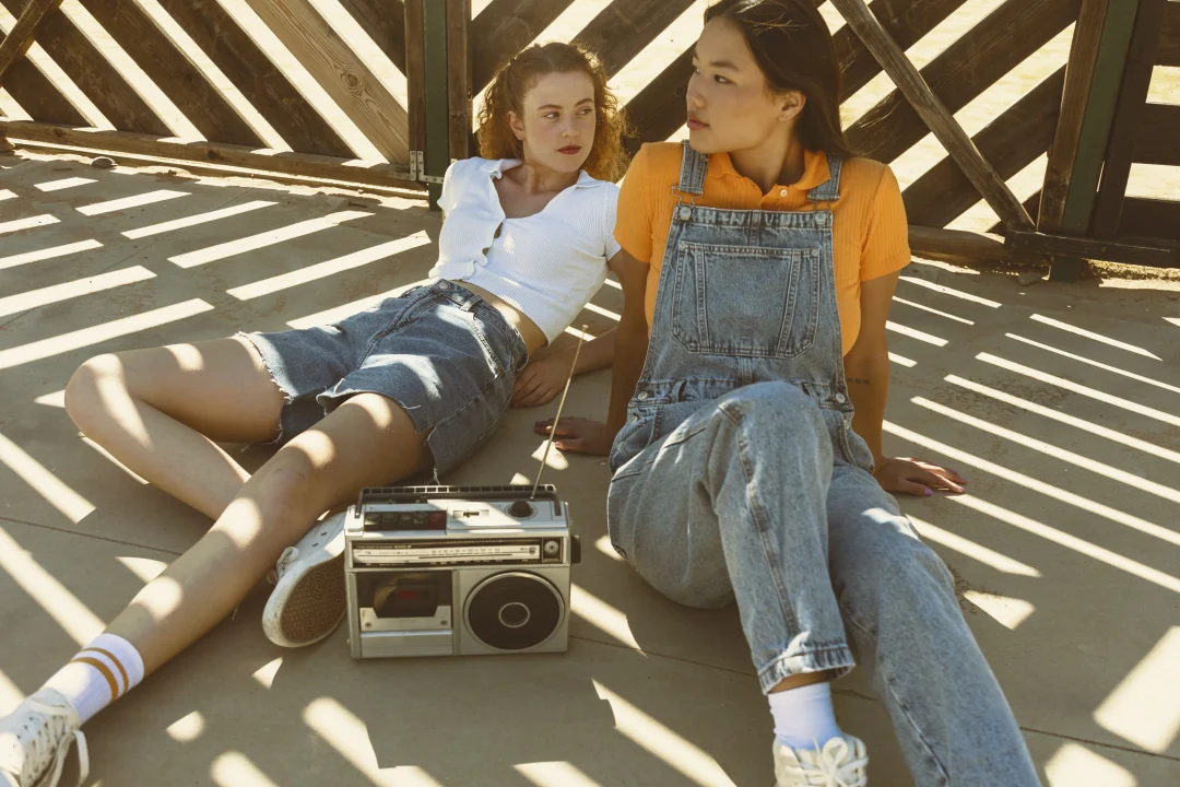 Zwei junge Frauen im Y2K Look mit Jeans-Shorts und Latzhose sitzen entspannt mit einem Retro-Kassettenrekorder in der Sonne.