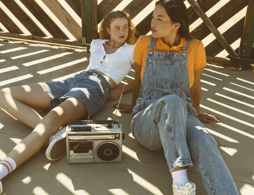 Zwei junge Frauen im Y2K Look mit Jeans-Shorts und Latzhose sitzen entspannt mit einem Retro-Kassettenrekorder in der Sonne.