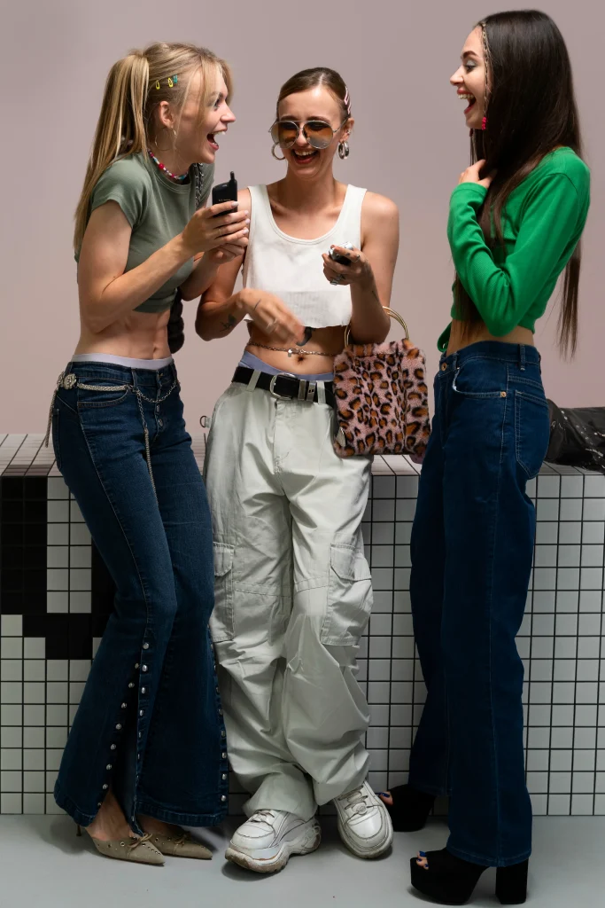 Drei junge Frauen im Y2K Look mit Hüfthosen, Cargo Pants, Crop Tops und Retro-Handys, die gemeinsam lachen.