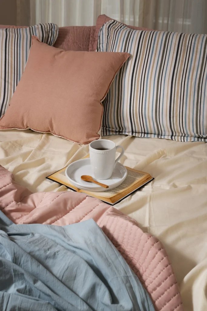 Gemütlich bezogenes Bett mit gemusterter und einfarbiger Bettwäsche sowie einer Kaffeetasse auf einem offenen Buch.