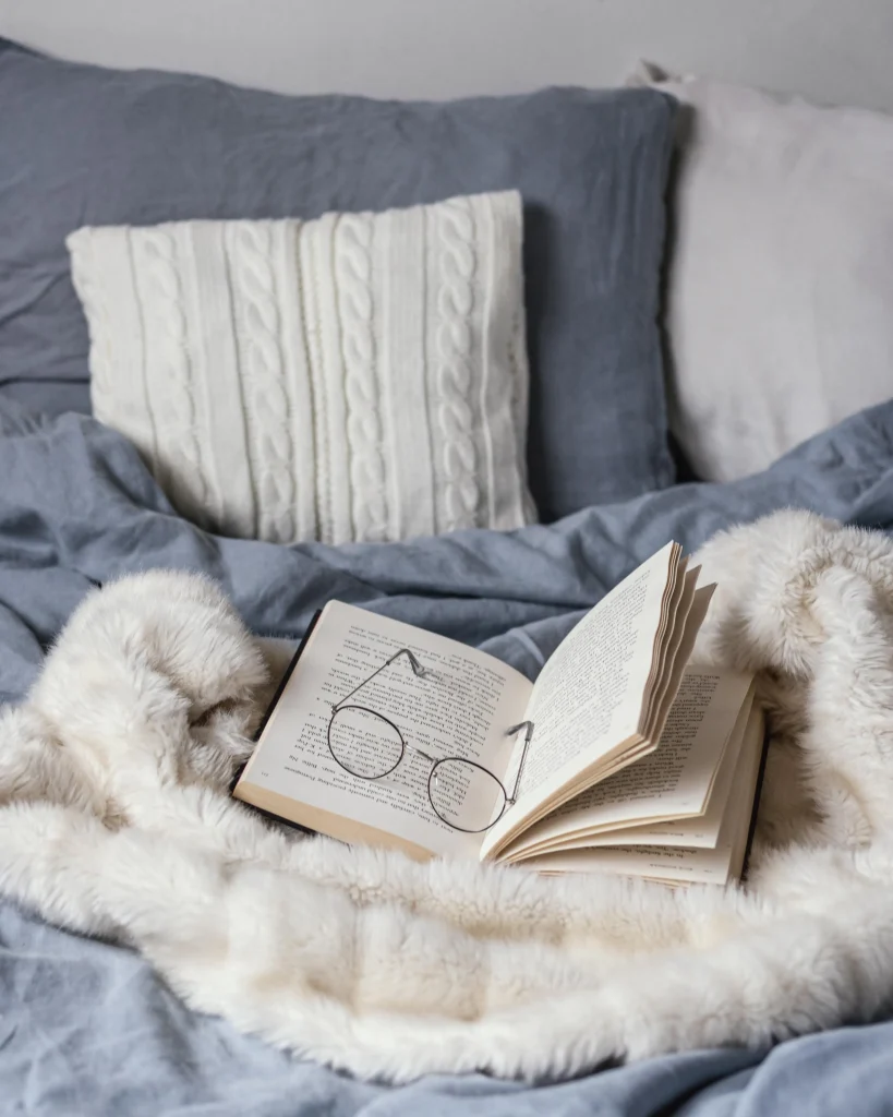 Offenes Buch mit Brille auf einem Bett mit grauer Bettwäsche und einer cremefarbenen Felldecke.