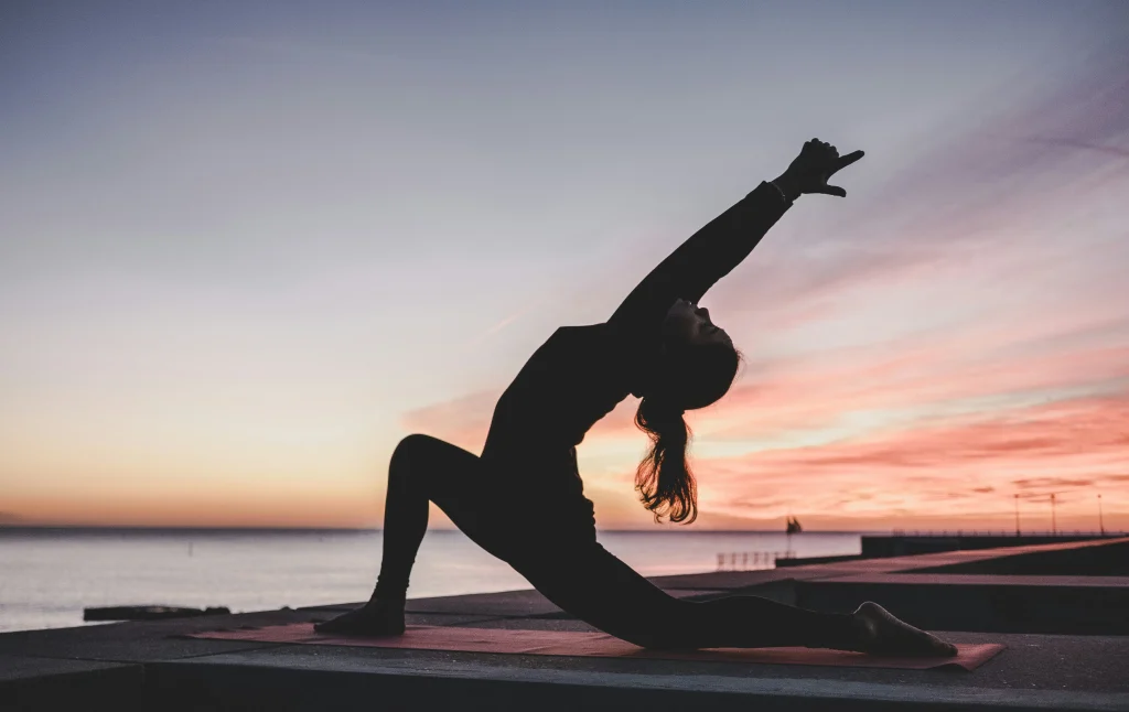 Frau in einer Hatha-Yoga-Pose bei Sonnenuntergang am Meer – Silhouette in tiefer Ausfallschritt-Haltung