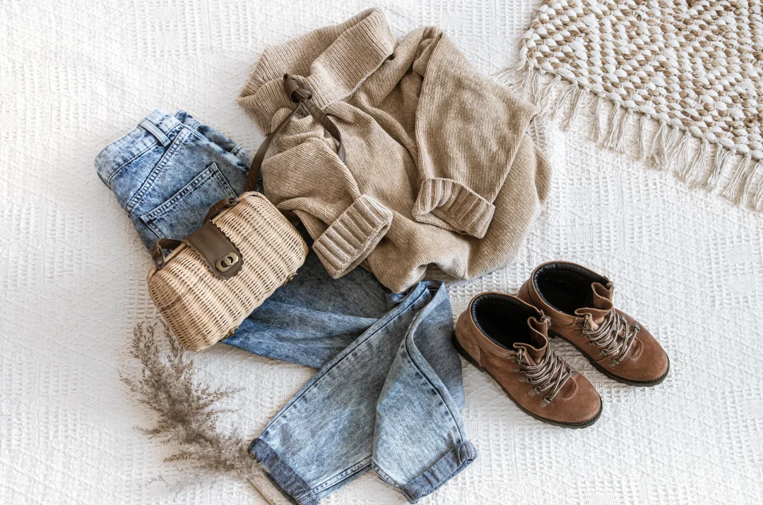 Capsule Wardrobe mit Jeans, beigem Strickpullover, braunen Lederstiefeletten und Korbtasche auf hellem Stoffuntergrund.