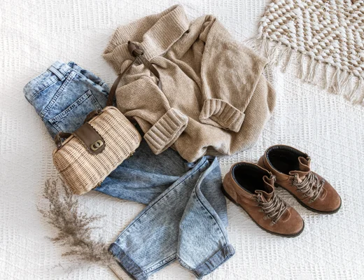 Capsule Wardrobe mit Jeans, beigem Strickpullover, braunen Lederstiefeletten und Korbtasche auf hellem Stoffuntergrund.