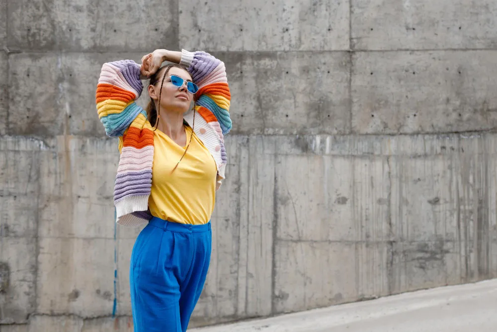 Frau mit buntem Pullover und blauer Hose im Dopamine Dressing Stil vor Betonwand