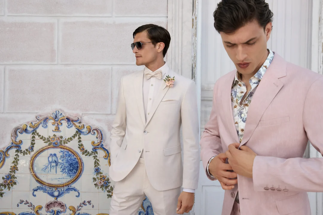 Zwei Herren in stylishen Hochzeitsanzügen, einer in Weiß, der andere in Rosa, stehen vor einer Wand mit floralen Kacheln.