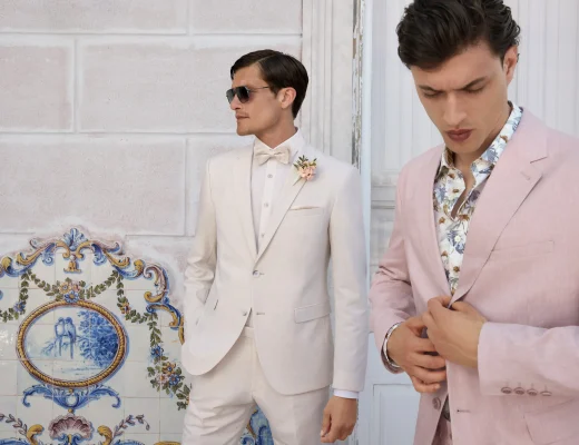Zwei Herren in stylishen Hochzeitsanzügen, einer in Weiß, der andere in Rosa, stehen vor einer Wand mit floralen Kacheln.