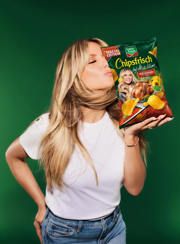 Heidi Klum posiert mit einer Packung funny-frisch Chipsfrisch Brath&auml;hnchen Style vor gr&uuml;nem Hintergrund.