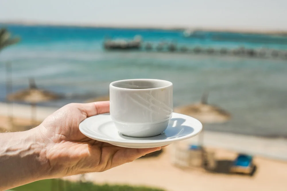 Hand hält eine weiße Kaffeetasse vor einem sonnigen Strand und türkisfarbenem Meer
