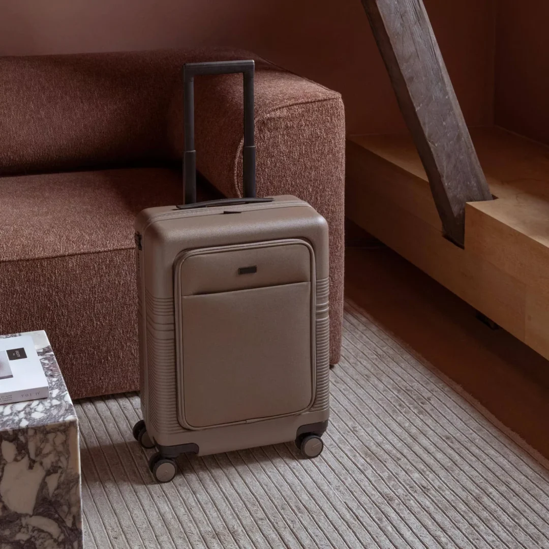 Beiger Koffer mit Vordertasche steht auf einem gestreiften Teppich vor einem braunen Sofa.