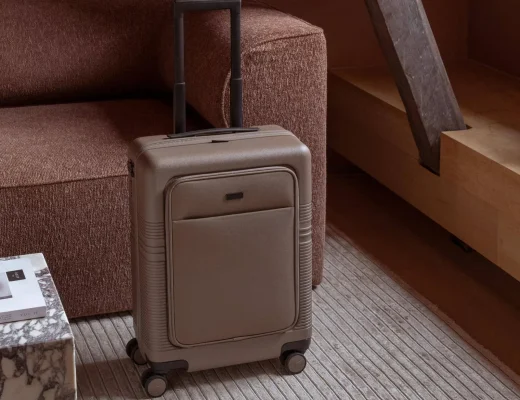 Beiger Koffer mit Vordertasche steht auf einem gestreiften Teppich vor einem braunen Sofa.