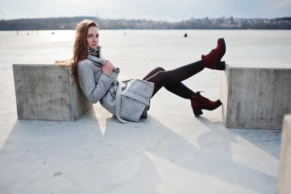 Frau mit roten Stiefeletten, grauem Mantel und schwarzer Strumpfhose am Wasser