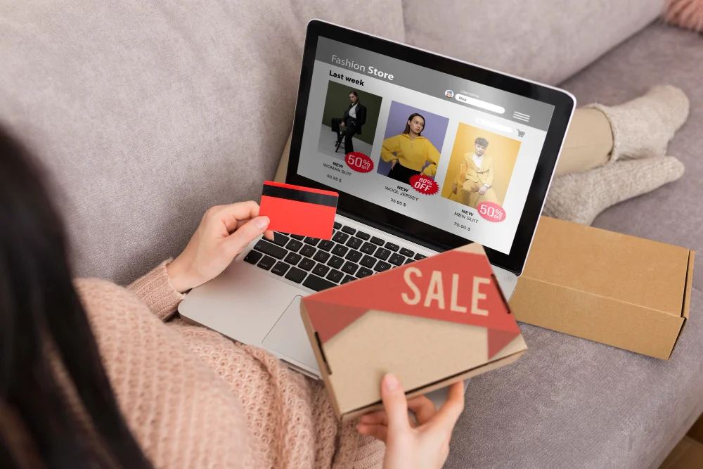 Frau kauft Kleidung in einem Online-Modegeschäft am Laptop und hält eine Kreditkarte sowie ein Paket mit Sale-Aufdruck.