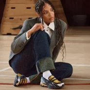 GANNI x New Balance 1906L in Fall Leaf/Black/Silver Metallic – Sneaker-Loafer mit Riemendetail und Metallic-Overlays, gestylt zu Blazer, weißem Hemd und Wide-Leg-Denim in einer Sporthalle.