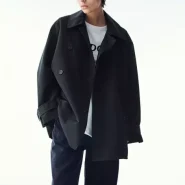 Damen Trenchcoat kurz in Schwarz, oversized, doppellagige Knopfleiste, über weißem T-Shirt und dunkler, weiter Hose im Studio.