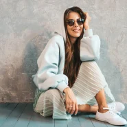 Casual chic Look: Frau mit pastellfarbenem Hoodie, plissiertem Midirock und weißen Sneakern, sitzend vor grauer Betonwand, mit Sonnenbrille