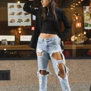 Streetstyle mit Ripped Jeans: Frau in hellblauer, stark zerrissener Jeans, schwarzem Top, Blazer und Cap vor einem Restaurant.