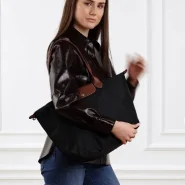 Frau trägt eine schwarze Longchamp Le Pliage Tote mit braunen Lederhenkeln über der Schulter; dunkles Hemd, Jeans, weißer Wandhintergrund.