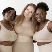 Drei Frauen verschiedener Körperformen tragen hautfarbene Shapewear von Creamy Fabrics und stehen lächelnd Arm in Arm vor hellem Hintergrund.