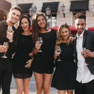 Gruppe junger Erwachsener in eleganter Dresscode-Cocktail-Kleidung, schwarze Midikleider und dunkler Blazer, mit Weingläsern auf einer Stadtterrasse am frühen Abend.