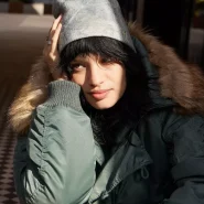 Person mit dunklem Haar trägt eine graue, leicht gemusterte Mütze und eine grüne Winterjacke mit Fellkapuze, während sie in der Sonne sitzt.