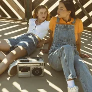 Zwei junge Frauen im Y2K Look mit Jeans-Shorts und Latzhose sitzen entspannt mit einem Retro-Kassettenrekorder in der Sonne.