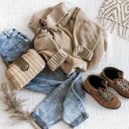 Capsule Wardrobe mit Jeans, beigem Strickpullover, braunen Lederstiefeletten und Korbtasche auf hellem Stoffuntergrund.