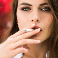 Nahaufnahme einer Frau mit grünen Augen und dunklem Smokey-Eye, die eine Zigarette raucht.